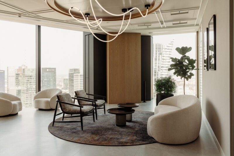Modern office lounge at the Baker McKenzie Warsaw office with cream chairs, circular tables, city views, indoor plants, and a sculptural ceiling light fixture.