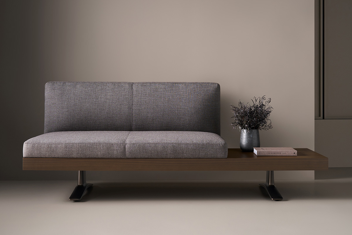 A modern sofa with grey cushions, a wooden base, and metal legs; a small side surface holds a metal vase with dried flowers and two books.