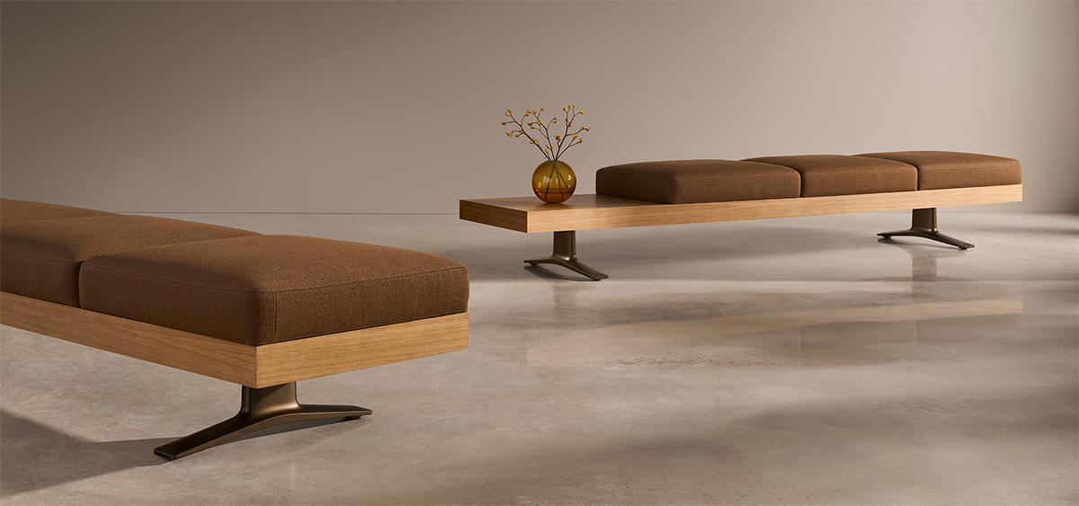 Two modern wooden benches with brown cushions on metal legs, placed on a polished floor. One bench has a small brown vase with branches on it. Neutral wall in the background.