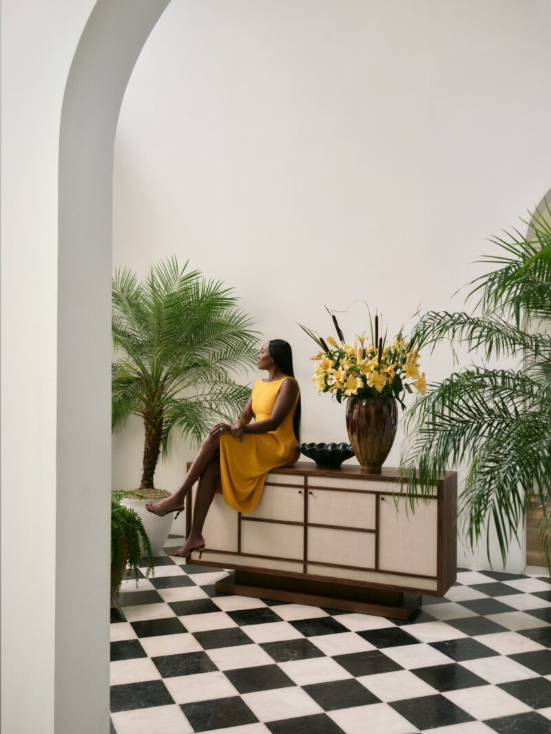 A woman in a yellow dress sits on a Crate & Barrel sideboard in a room with black and white checkered floor, potted plants, and a tall flower arrangement—a setting reminiscent of the modern elegance often inspired by Venus Williams.