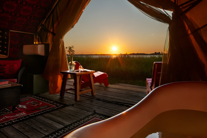 A sunset view over a lake seen from inside an Urban Cowboy Tent, with a bathtub, red chairs, a small table, and patterned rugs on a wooden floor.