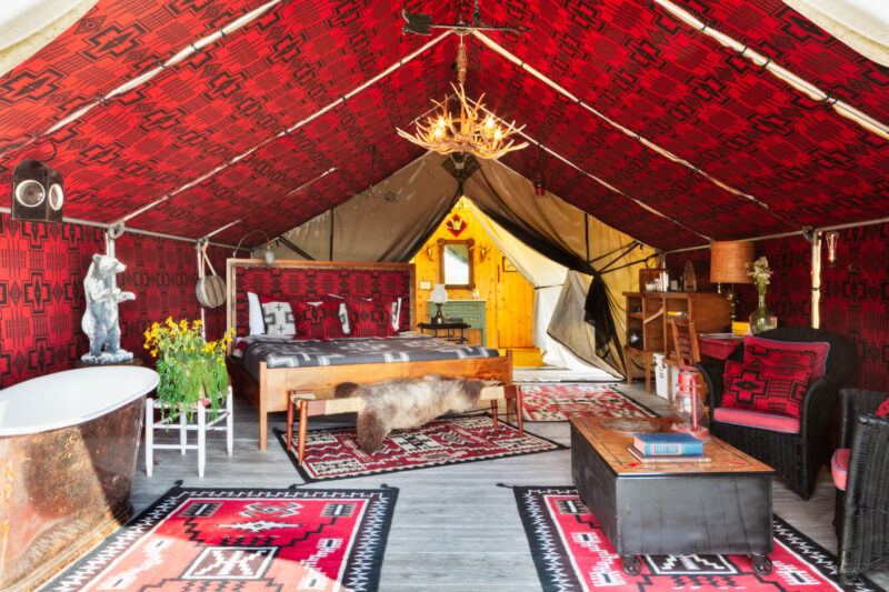 A luxury Urban Cowboy Tent interior with red patterned decor, a bed, wooden furniture, a freestanding tub, a chandelier, and rugs on a wooden floor.