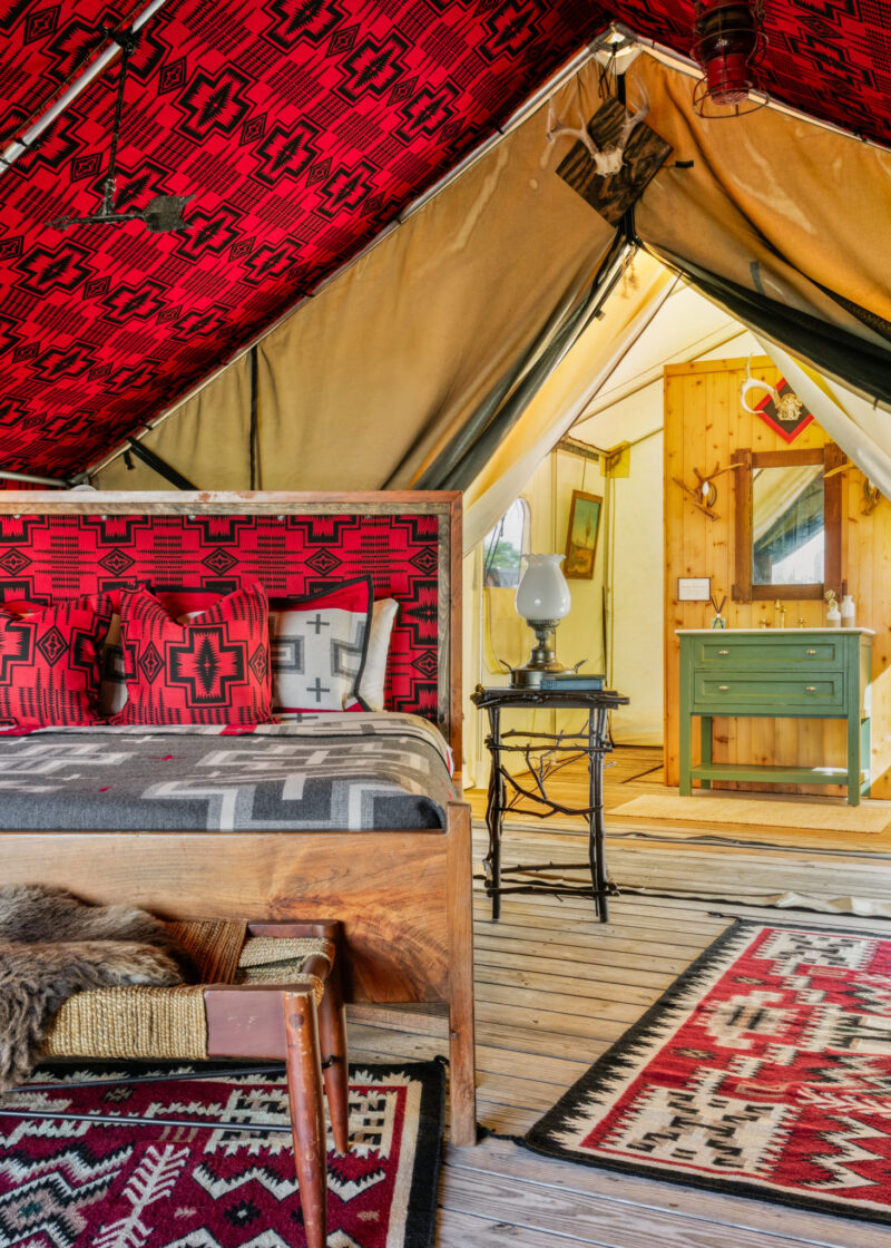 A glamping tent interior, known as the Urban Cowboy Tent, features a red patterned bed, wooden furniture, rugs, and a small green vanity in the back room.