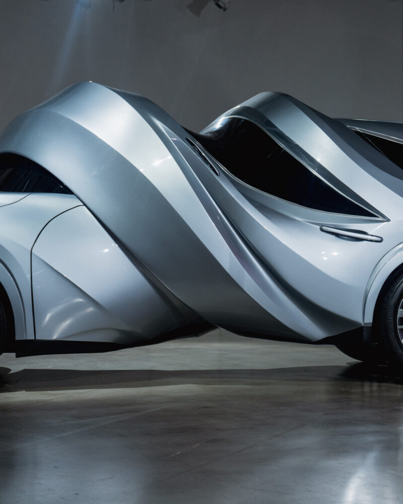 Futuristic silver concept car with a twisted, sculptural body design, reminiscent of the MSCHF Lexus MOTOMORPHOSIS, displayed indoors on a concrete floor.