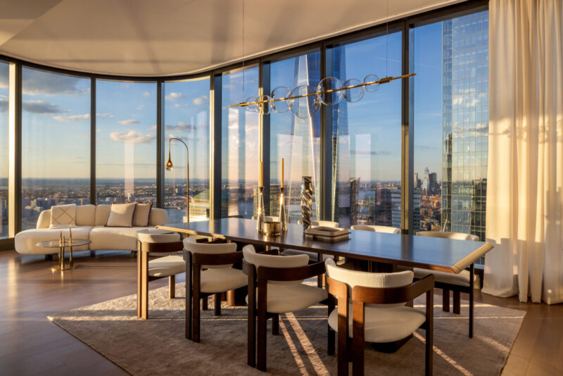 Modern dining room with a wooden table, cushioned chairs, and floor-to-ceiling windows showing a cityscape at sunset. A white sofa and gold accents sit near the curved glass wall.