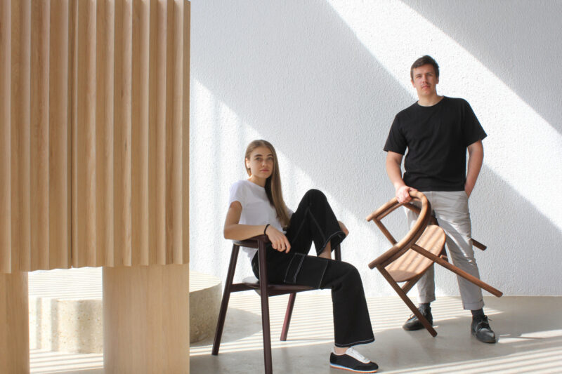A woman sits on a chair with her legs up, and a man stands holding a chair in a minimalist room with sunlight streaming through.