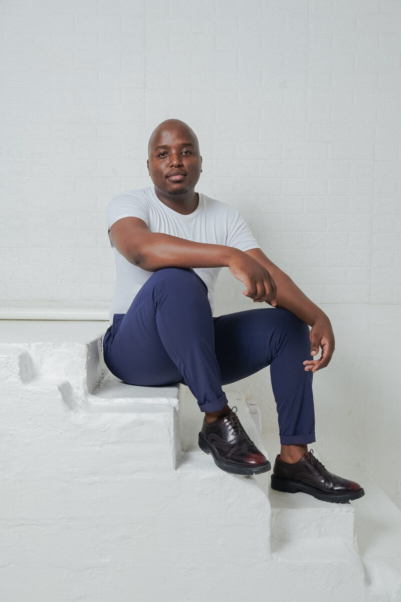 A man in a white t-shirt and blue pants sits on white stairs, resting one arm on his knee and looking at the camera.