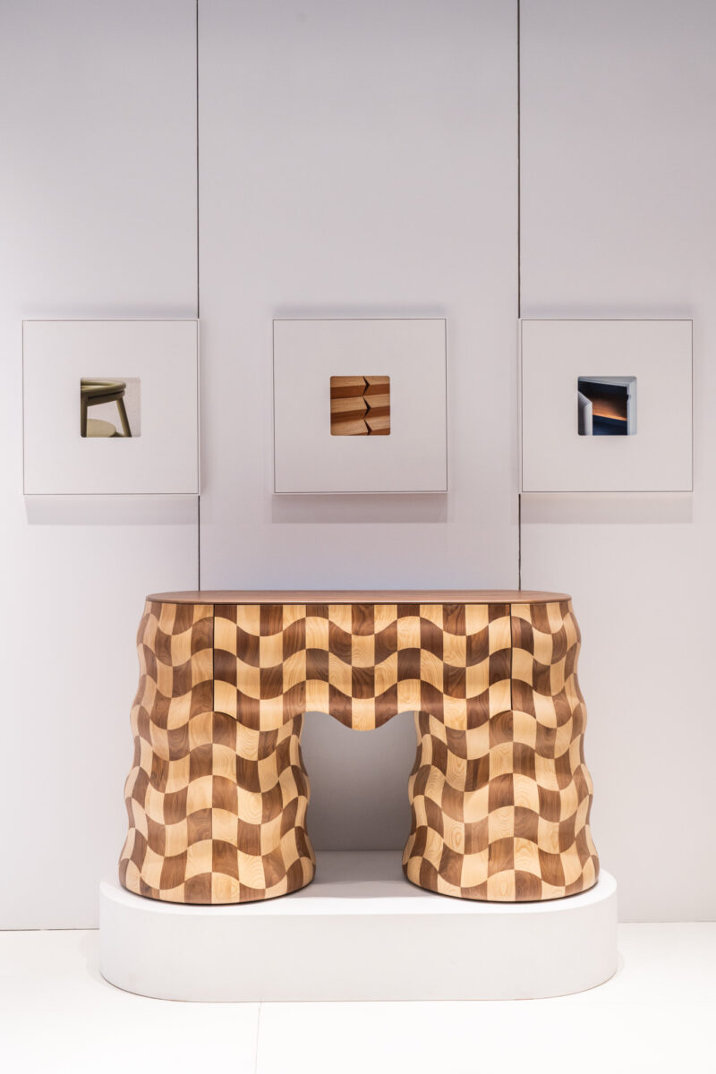 A wavy, checkered wooden table is displayed on a white platform with three minimalist framed artworks hanging on the white wall behind it.