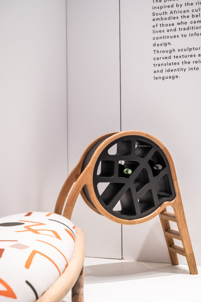 A modern wooden and black circular sculptural chair sits next to a patterned cushioned bench against a white wall with partial text visible.