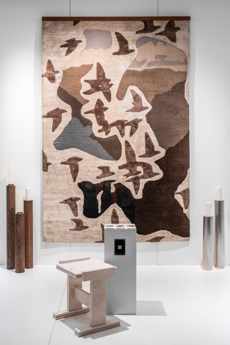 A large abstract wall tapestry with bird-like shapes hangs above minimalist wooden and stone furniture and cylindrical sculptures in a modern gallery space.