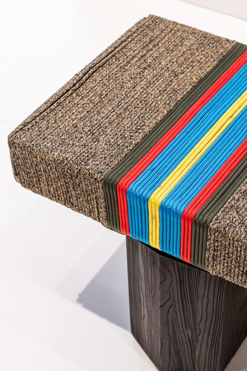 Square stool with a textured brown cushion, featuring blue, red, yellow, and olive green stripes on top, supported by a black wood-patterned base.