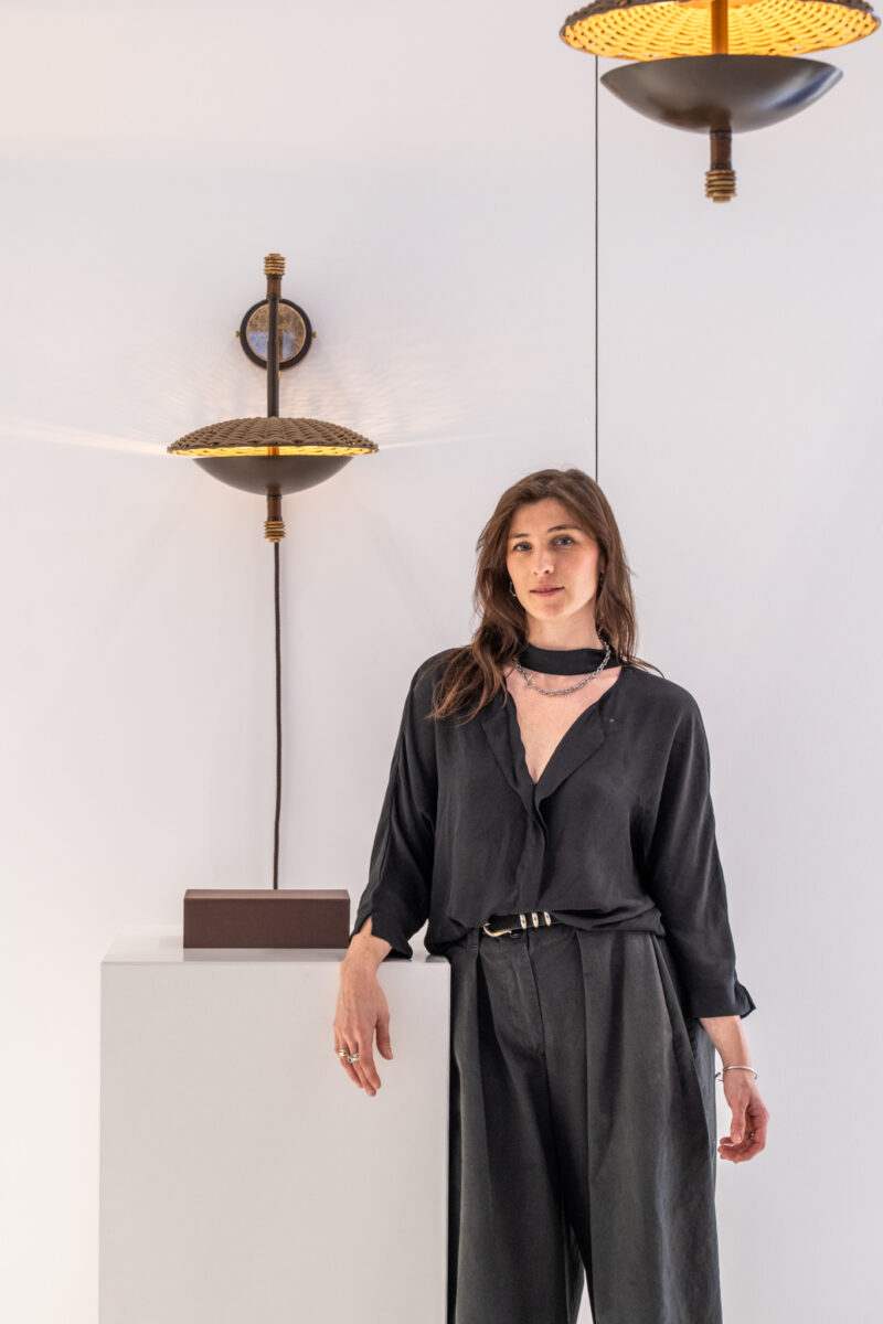 A woman in dark clothing stands beside a white pedestal with a modern wall lamp and pendant light on a plain background.