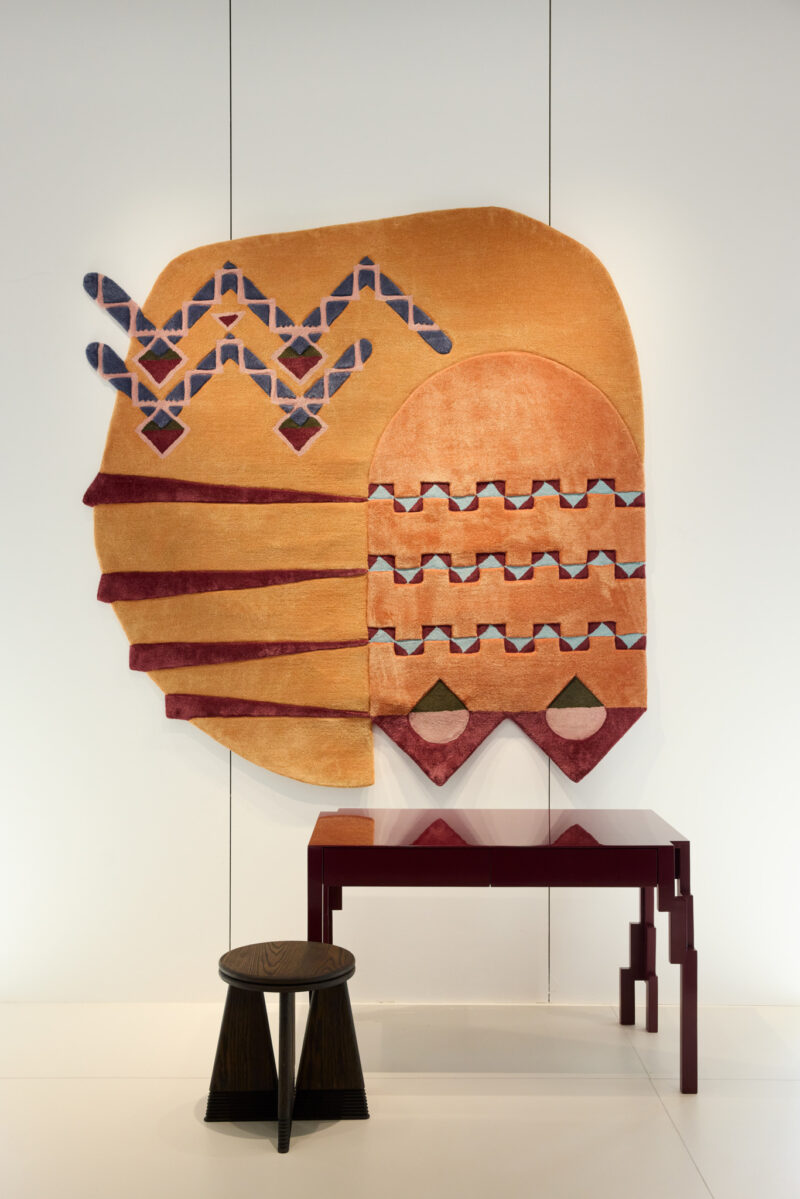Geometric-patterned wall tapestry in warm tones hangs above a modern maroon desk and a small dark wooden stool in a minimal white room.