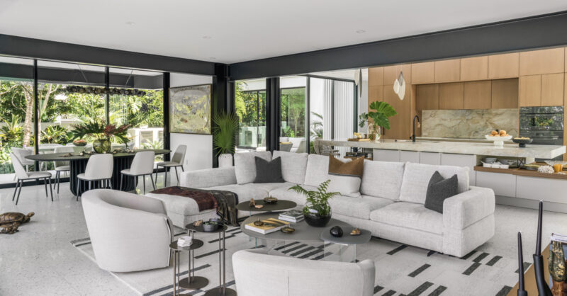 Modern open-plan living space inspired by Paul Rudolph Biggs Residence, with a white sectional sofa, two armchairs, dining table, and garden views through large windows.