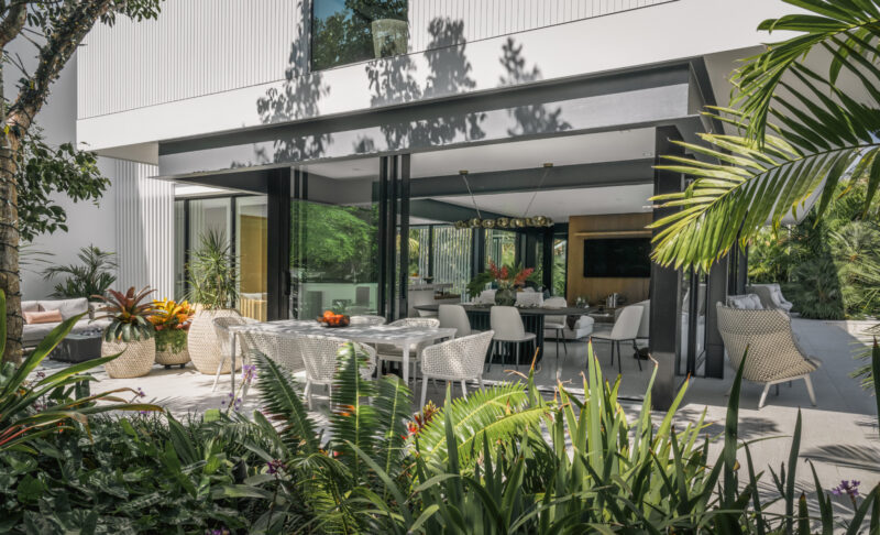 Modern patio with white outdoor furniture, a dining table, and lush green plants, adjacent to the Paul Rudolph Biggs Residence with large glass doors.