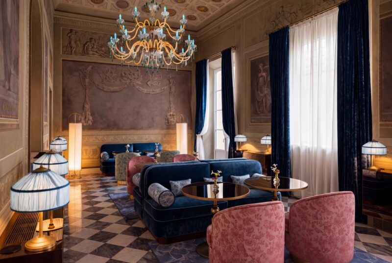 A lounge at Palazzo Sogni Florence with blue velvet sofas, pink patterned armchairs, small circular tables, and a large chandelier in a room with a checkerboard floor and dark blue curtains.