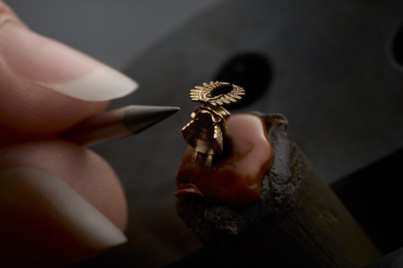 A metalworker’s tool points toward the gold laurel wreath emblem of the Roger Dubuis Excalibur, mounted on a dark, wax-like base.