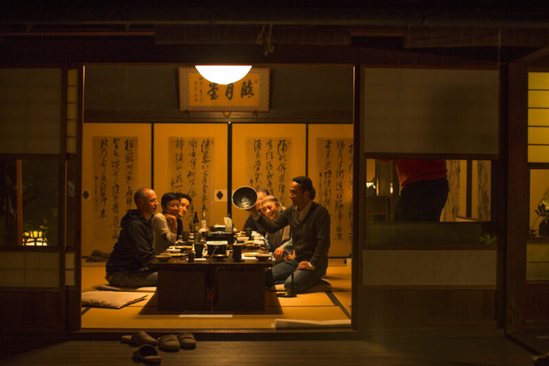 A group of people sit on floor cushions around a low table in a room with sliding paper doors, as one man holds up a bowl.