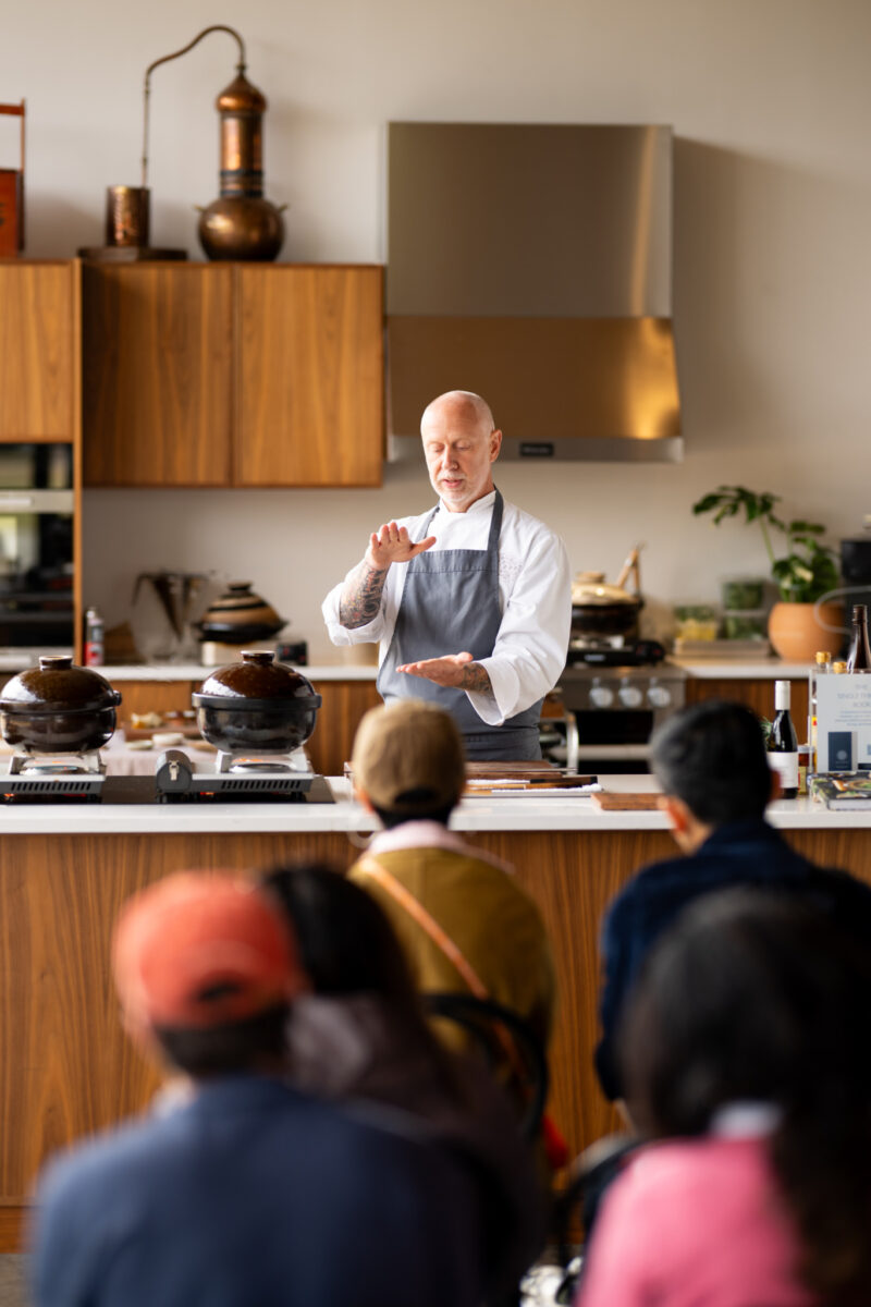 A chef in a white uniform and grey apron demonstrates cooking techniques to a small audience in a kitchen.