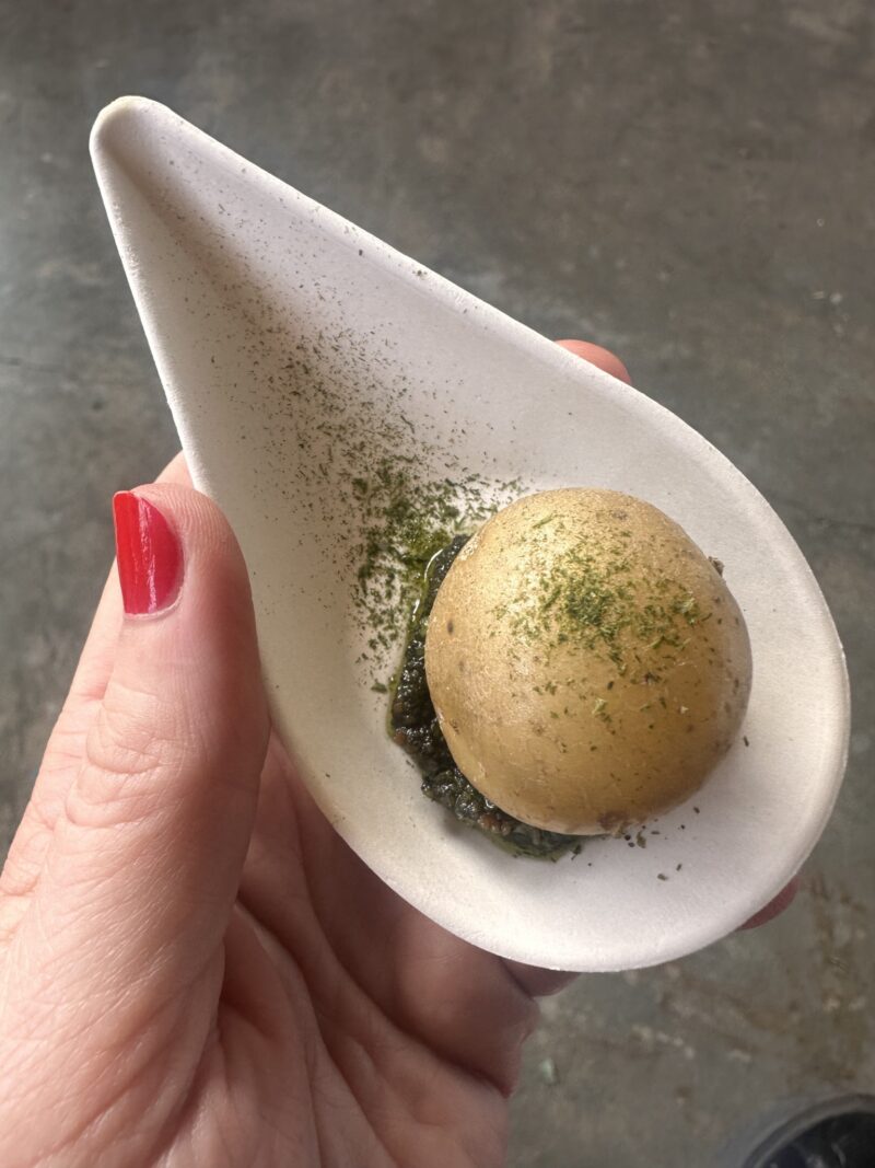 A small boiled potato on a bed of pesto, sprinkled with green herbs, served on a white tasting spoon.
