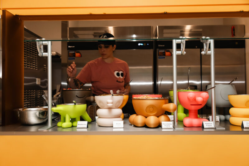 A server in a pink shirt scoops food from a counter at Softville NYC, filled with colorful, character-shaped bowls labeled with flavor names.