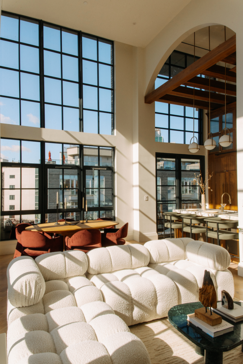 Modern living room with a white sectional sofa, large grid windows, views of city buildings, wooden dining table with red chairs, and an open kitchen under exposed wooden beams.