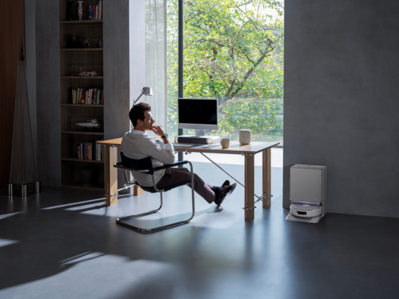 A man sits at a desk facing a computer monitor in a room with a white Narwal Flow 2 robotic vacuum cleaner parked on the floor nearby.