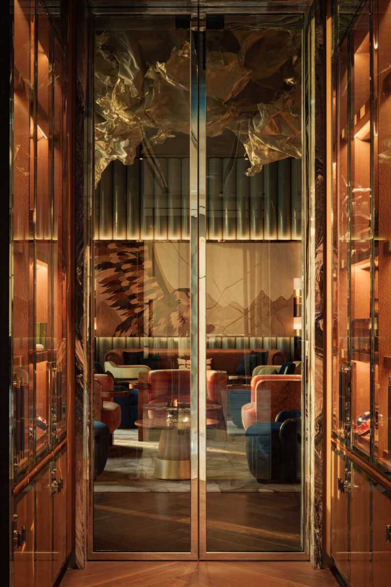 View through glass double doors into a lounge area with upholstered chairs, a low coffee table, and a mural of a bird on the wall.