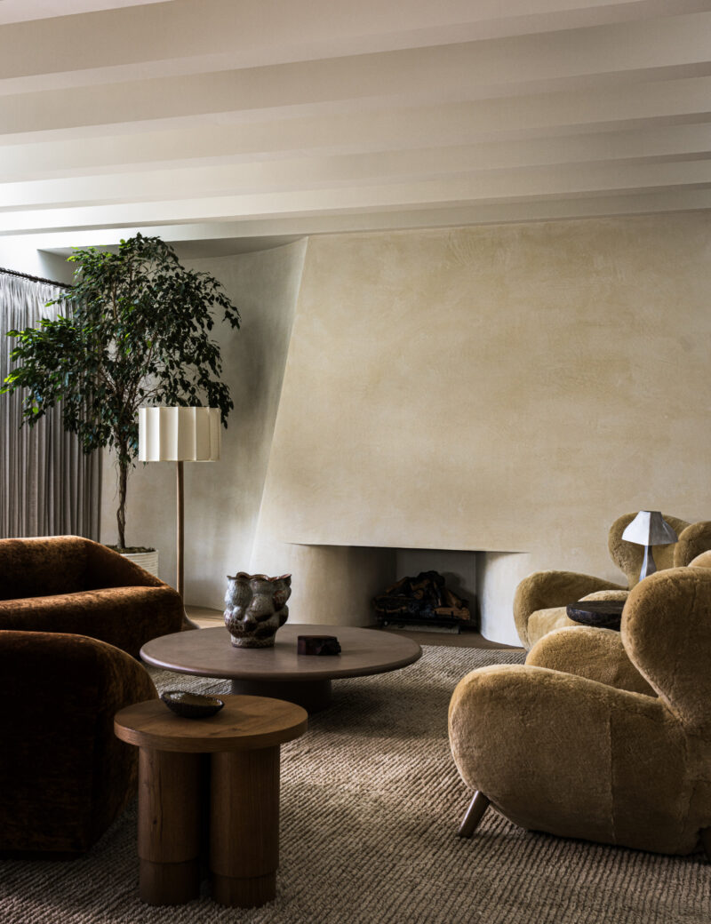 Living room with a large, beige fireplace, curved armchairs, a round coffee table, and a floor lamp next to a large potted plant.