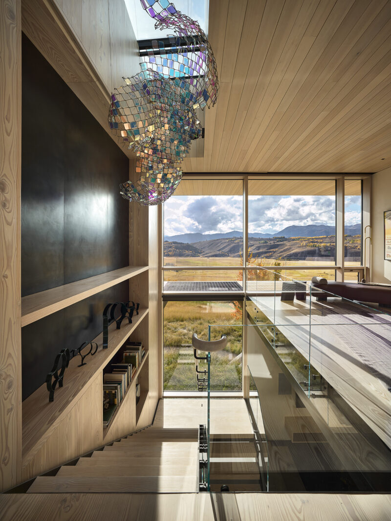 Modern interior with wooden staircase, built-in shelving, a large iridescent sculpture hanging from the ceiling, and a floor-to-ceiling window overlooking a mountain landscape.