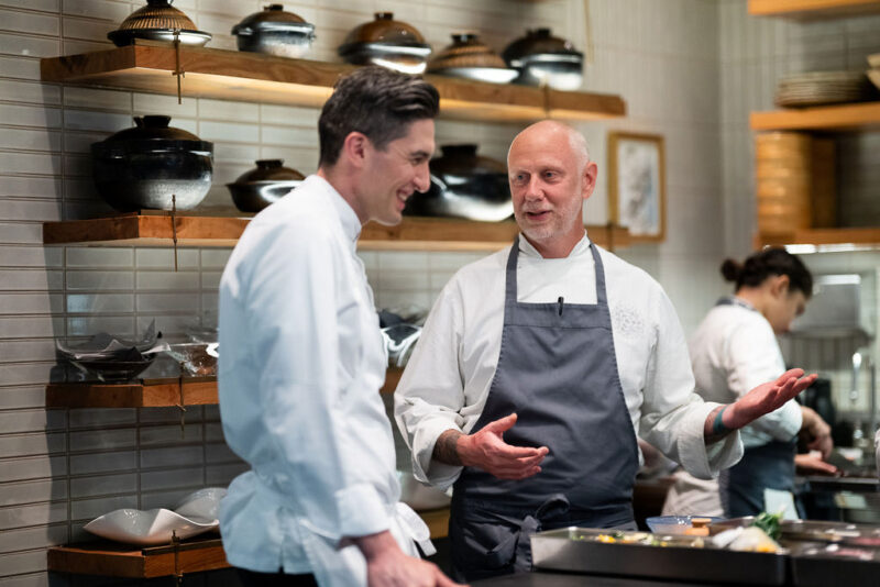 Two chefs in white uniforms talk in a kitchen, with one gesturing toward a workspace.
