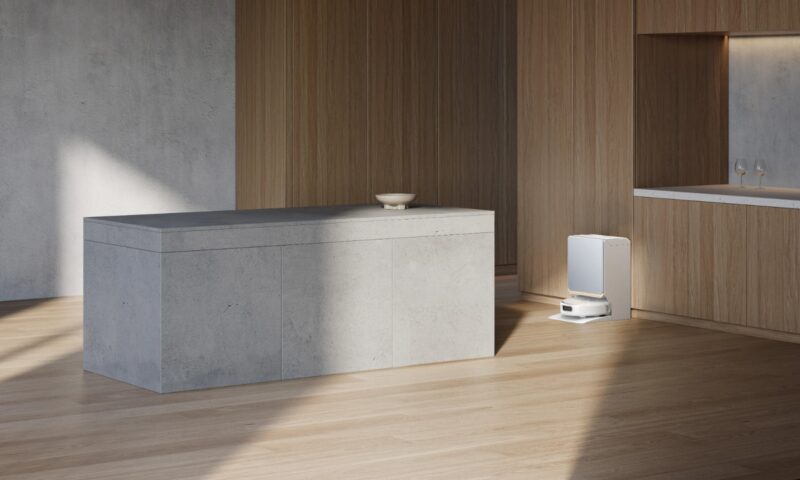 A white Narwal Flow 2 robot vacuum docked at its base station in a modern kitchen with a gray island and wood cabinetry.
