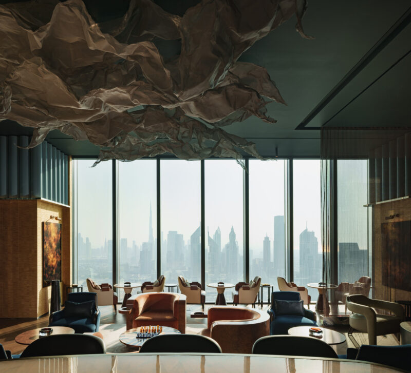 Modern lounge overlooking a bright city skyline through floor-to-ceiling windows, featuring multiple seating areas and a large sculptural installation hanging from the ceiling.