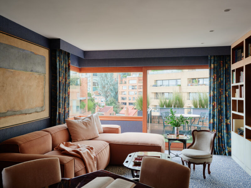 A living room with an orange sectional sofa, a patterned armchair, a built-in bookshelf, and a large window overlooking a balcony and city buildings.