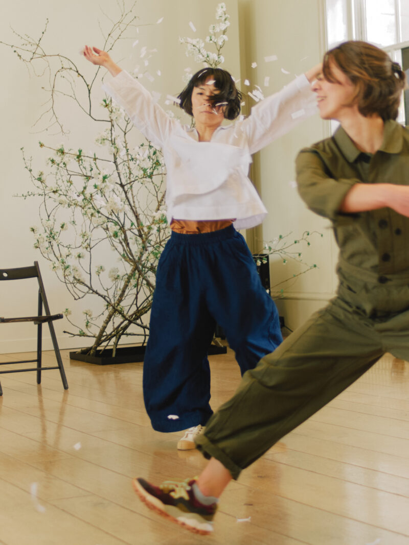 Two people dance energetically indoors; one throws white confetti with arms raised, while Suzie de Rohan Willner moves in the foreground. Sparse green branches adorn the background.