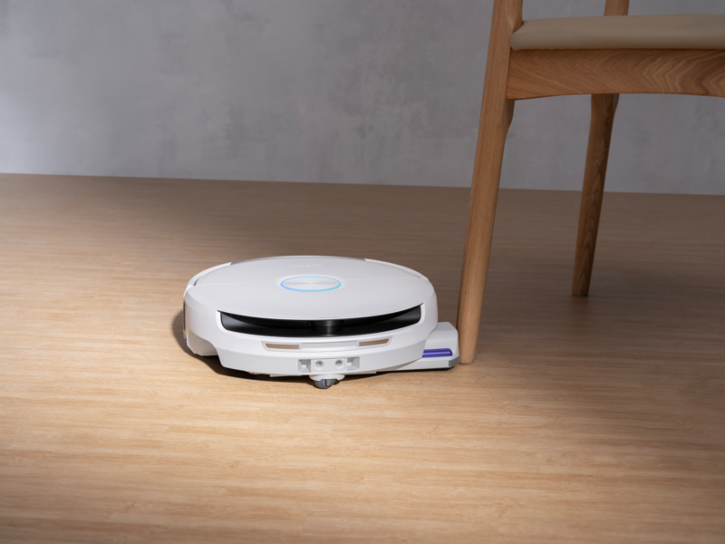 A white robot vacuum cleaning near the leg of a wooden chair.