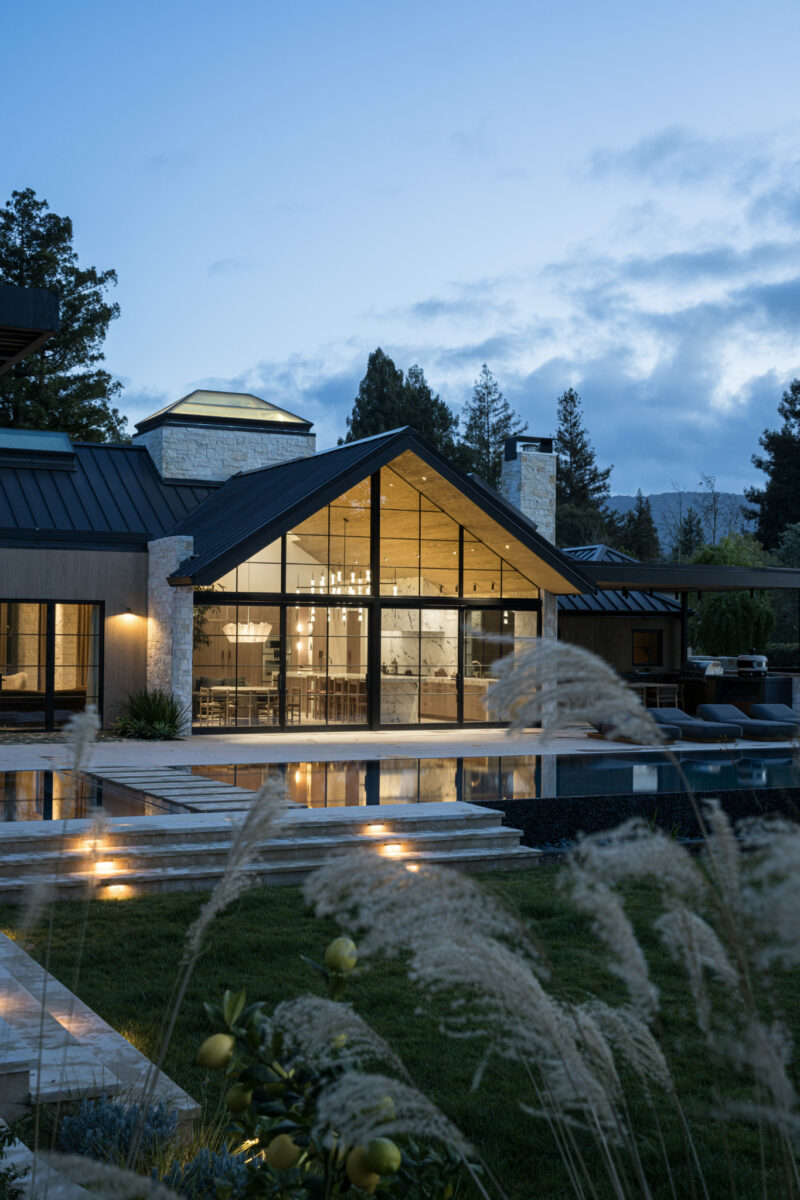 Modern house with large glass windows overlooking a swimming pool at dusk.