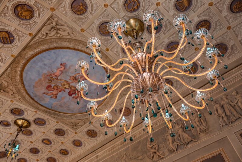 An ornate chandelier hangs below a coffered ceiling at Palazzo Sogni Florence, decorated with zodiac motifs and a circular fresco of cherubs.