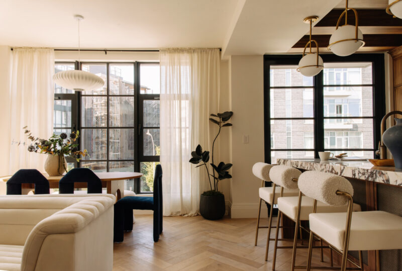 Modern living and dining area with large windows, light wood floors, a round dining table, upholstered chairs, a marble kitchen island, and potted plants. Natural light fills the room.