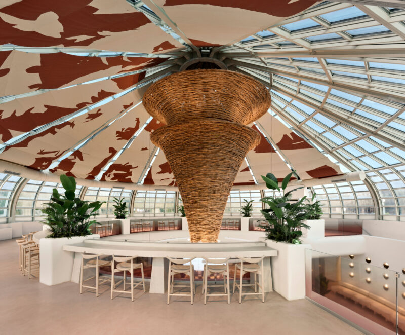 A curved bar with wooden stools beneath a large, woven, cone-shaped pendant light in a Local People greenhouse-style cafe with a glass-paneled domed ceiling.