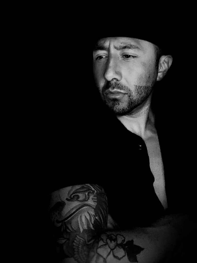 Black and white portrait of Alfonso Verduzco, a man with a tattooed arm, looking to the side.