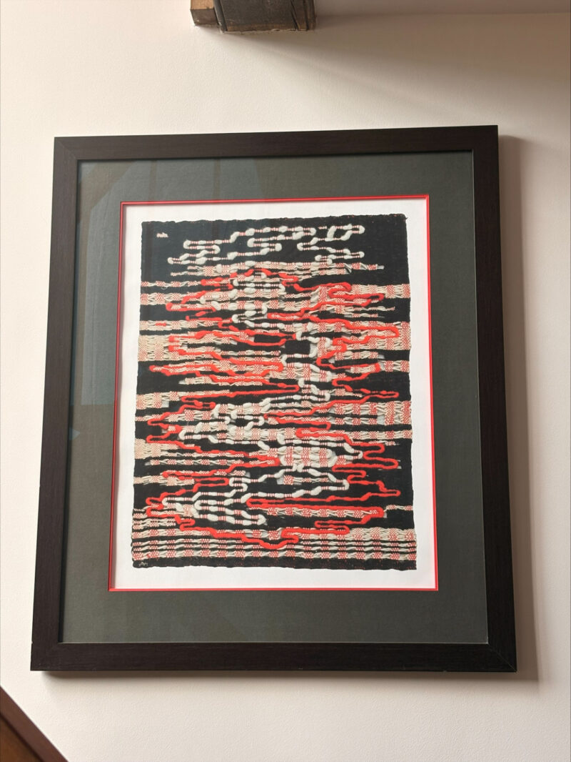 Framed textile artwork featuring abstract black, white, and red horizontal patterns, displayed against a white mat and dark frame on a wall.
