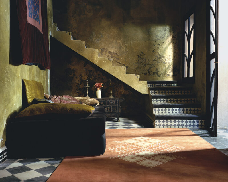 A living room with a low black sofa, a small side table holding a vase, a staircase with patterned tile risers, and a large orange rug from our designer rug collections featuring a geometric pattern.