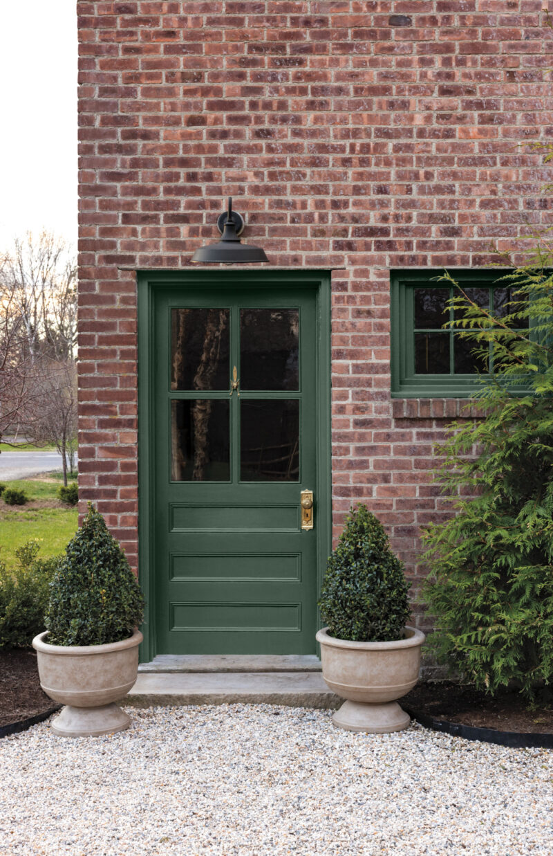 A green Benjamin Moore Color of the Year 2027 front door with a glass pane, set in a red brick wall with two potted topiaries on a gravel path.