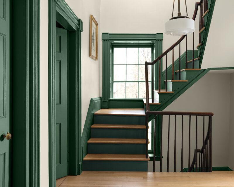 A staircase with wooden treads and dark green trim, finished in Benjamin Moore Color of the Year 2027, leading to a window with a matching green door and dark wooden railing.