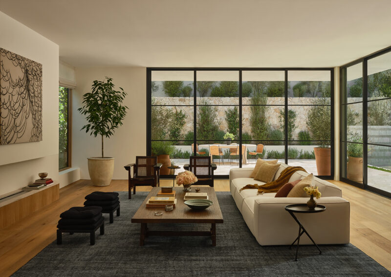 Modern living room with large windows, a white sofa, wooden coffee table, potted plant, and view of an outdoor patio with table and chairs.