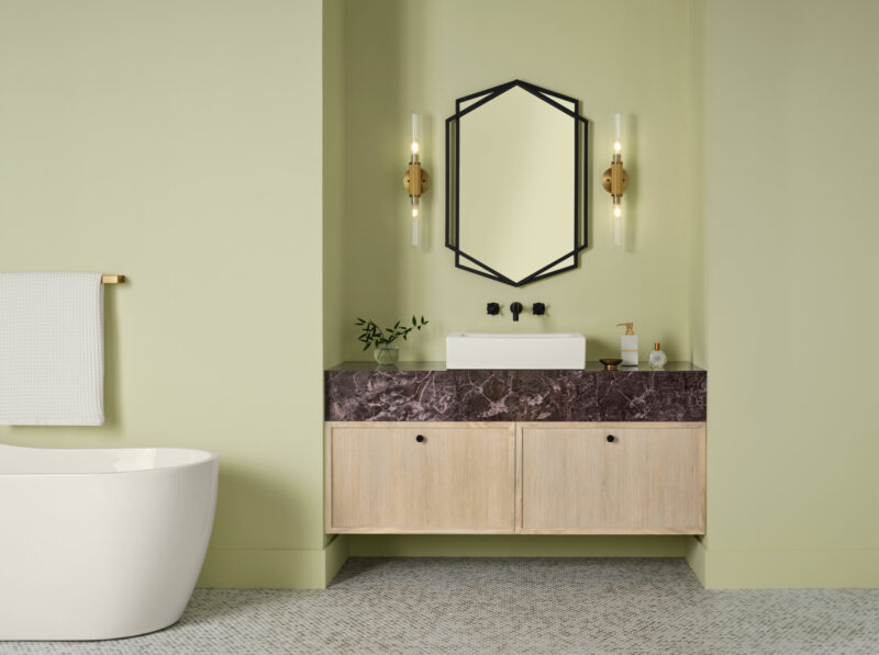 A white vessel sink on a marble countertop with wood cabinets sits under a hexagonal mirror between two wall sconces, styled in the Sherwin-Williams 2027 Color of the Year, near a freestanding tub.