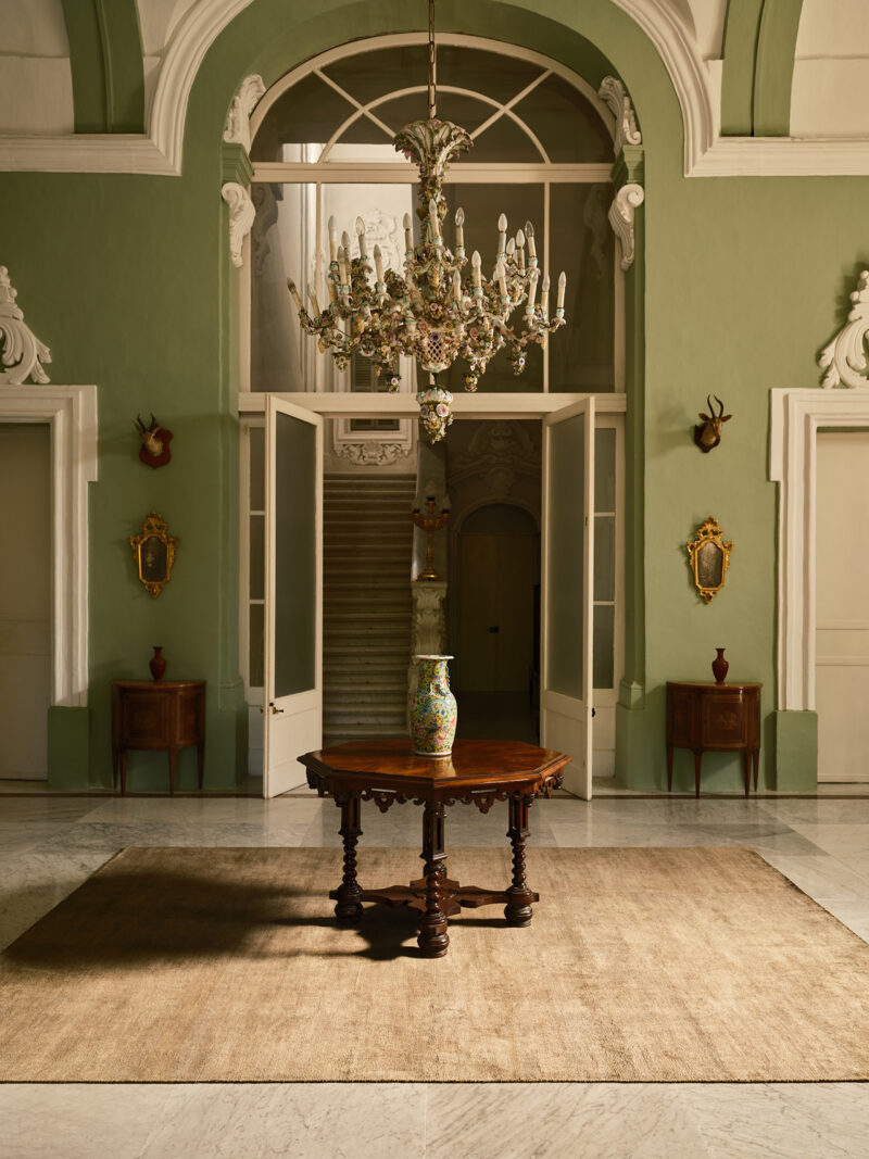 Elegant foyer with green walls, a large chandelier, a round wooden table with a vase, matching side tables, and open double doors leading to a staircase in the background.