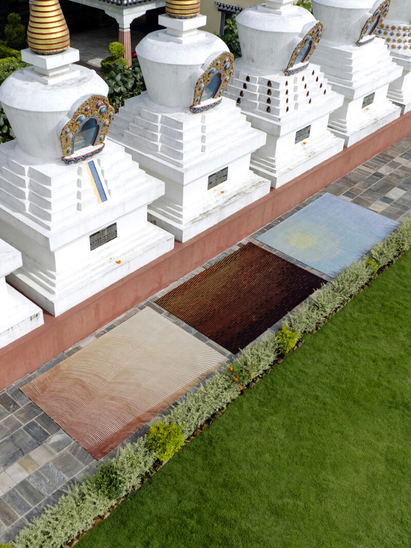 A row of white stupas stands behind three rectangular mats in different colors, laid on a stone walkway bordered by grass and shrubs.