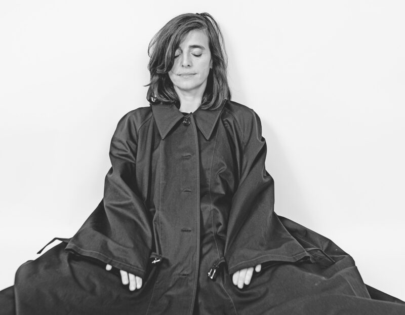 A person with their eyes closed sitting in a meditation pose, wearing a dark buttoned coat.